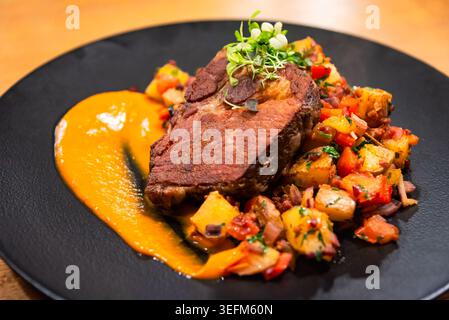 Hearty main course featuring roasted meat, colorful potato hash, and bright orange puree served on an elegant dark dish Stock Photo