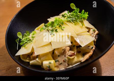 Close-up of delicious creamy pappardelle pasta with chicken, mushrooms, topped with shaved parmesan cheese and fresh microgreens, served in a black bo Stock Photo