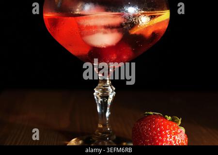 Red cocktail with ice, lemon slice and strawberries in a chilled glass, vibrant and refreshing on a dark backgroundperfect for summer parties, bars, m Stock Photo