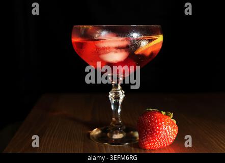 Cocktail glass with red chilled drink, ice and citrus cherry garnish on a wooden table beside a fresh strawberry, studio still life evoking summer ref Stock Photo
