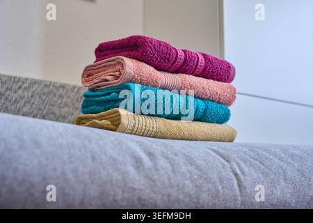 A stack of four neatly folded colorful bath towels in magenta, pink, teal, and yellow resting on a grey sofa Stock Photo