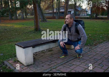 Middle-aged man crouching beside a park bench while wearing a large backpack and a grey fleece jacket, gazing sideways with a pensive and curious expr Stock Photo
