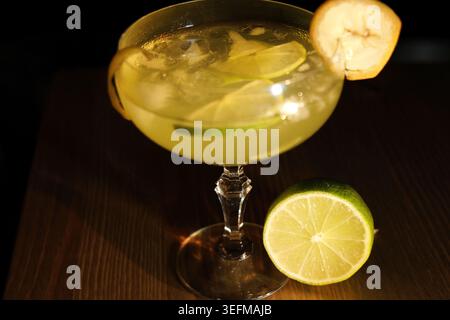 Refreshing alcoholic drink served in a coupe glass, garnished with lime slices, an orange peel twist, and a banana chip, sitting next to a cut lime on Stock Photo
