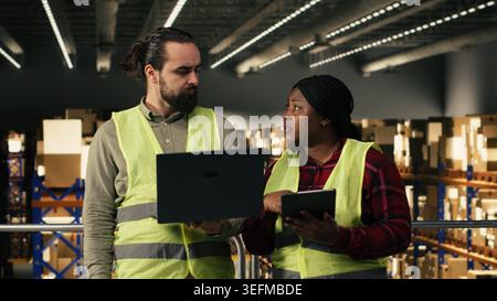 Multiethnic team reviewing shipping details and parcel tracking information on laptop, working together on platform. Organized fulfillment center environment reflects efficient package handling. Stock Photo