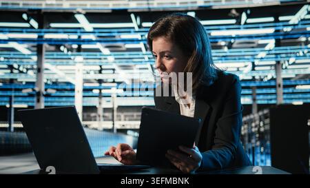 Data center software developer optimizing equipment rigs using devices. Woman in server farm running programming script on laptop and tablet, adjusting system parameters Stock Photo