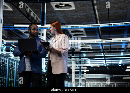 Data center engineering team monitoring system performance, identifying bottlenecks. Server farm teamworking colleagues using devices to implement optimizations improving efficiency Stock Photo