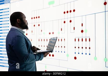 RD engineer oversees microprocessor plant production using laptop. African american man using notebook and digital wall display to analyze semiconductor fabrication steps, ensuring workflow efficiency Stock Photo