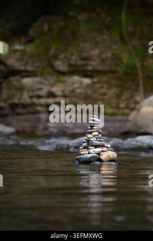 A unique stack of smooth stones, known as a cairn, balances harmoniously in still water, reflecting nature's beauty. Surrounded by lush greenery and g Stock Photo
