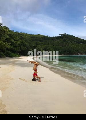 A woman performs an advanced headstand yoga pose on a pristine tropical beach. An elegant eagle-inspired variation with the legs Stock Photo