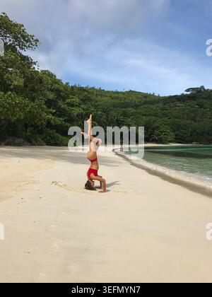 A woman performs an advanced headstand yoga pose on a pristine tropical beach. An elegant eagle-inspired variation with the legs Stock Photo