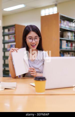 Young Asian woman with glasses holding notebook working on laptop computer at library desk with coffee cup smiling happy Stock Photo