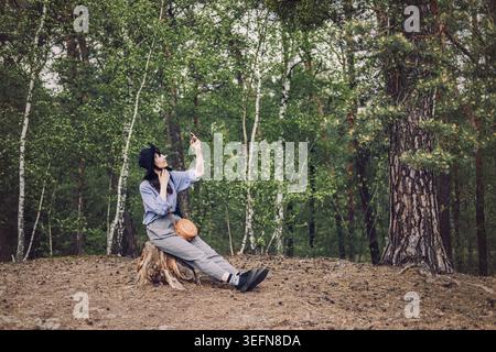 Person wearing hat sits on tree stump in forest and holds smartphone up toward trees. AI chatbot for everyday use, virtual assistant in nature Stock Photo