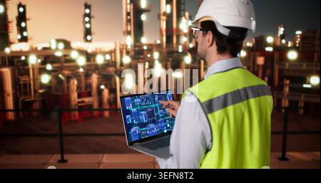 Engineer Monitors Refinery Data On Laptop While Inspecting Industrial Pipelines. Stock Photo
