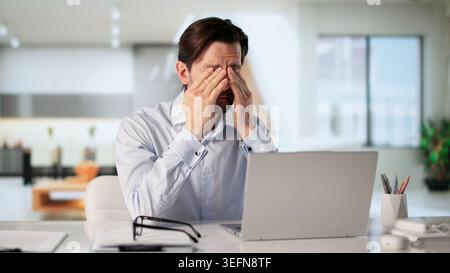 Tired Man Rubbing Sore Eye While Working On Laptop Feeling Stressed. Stock Photo