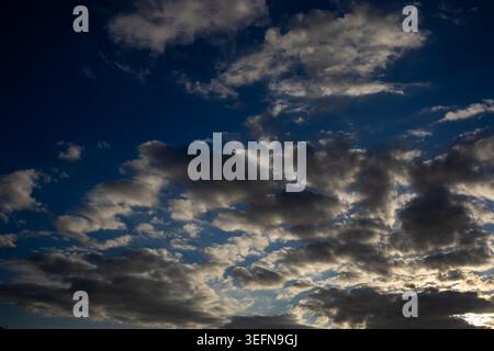 Broken cloud formations scattered across deep blue sky with warm light catching cloud bases from below Stock Photo