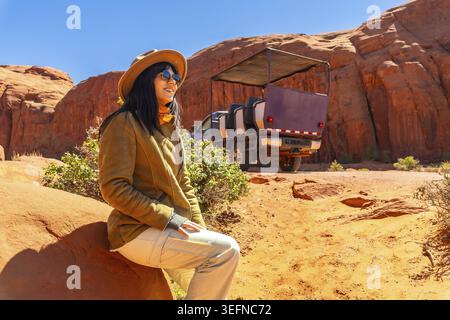 Woman sitting on a red rock, enjoying the scenic desert landscape of monument valley during a guided jeep tour, exploring the vast sandstone formation Stock Photo