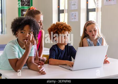 Diverse children leaning over silver laptop at classroom table, collaborating on school project Stock Photo