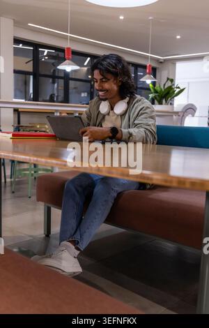 Indian man sitting at long wooden table in coworking space using tablet with headphones Stock Photo