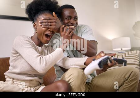 Happy, black couple and playing video games with surprise for fun competition, humor or comedy. Woman, console and playful man with eyes covered for Stock Photo
