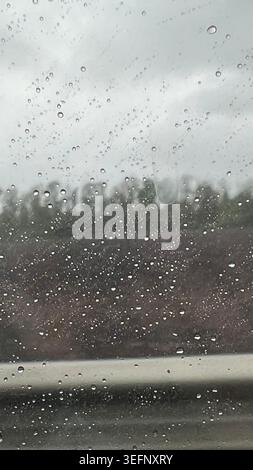 Raindrops close-up on a car window glass on a cloudy day with blurred forest and road background. Moody bad weather concept and safe driving in rain Stock Photo