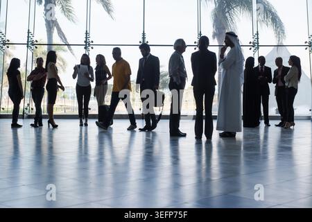 Silhouetted professionals gather in a modern glass atrium, conversing and waiting with luggage. A diverse mix of formal and casual dress suggests travel, networking, conferences and corporate meetings in a sunny setting. Stock Photo