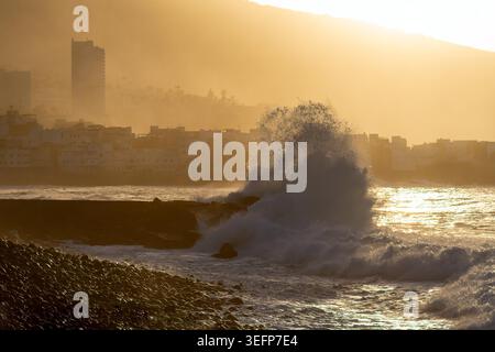 High sea waves on the background of the coastline in a haze. The waves beat against the stones on the shore. Spatter of waves, sea and pebbles shimmer with silver in the rays of the rising sun. Stock Photo