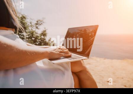 Laptop woman beach remote worker sits on a cliff and types on a computer while overlooking the vast ocean on a sunny day Stock Photo