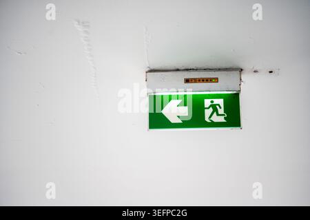 A green emergency exit sign with arrow and running figure is mounted on a white ceiling, providing clear guidance for safe evacuation in case of emerg Stock Photo
