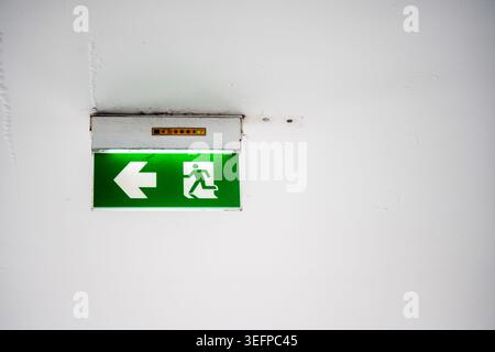 A green illuminated emergency exit sign with a left arrow and running figure, indicating the direction for evacuation in case of emergencies, mounted Stock Photo