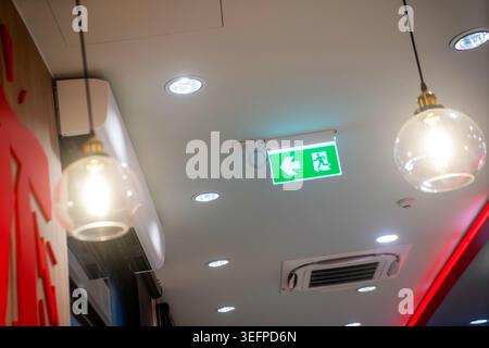 Modern indoor space showing a ceiling with a safety emergency exit sign illuminated in green, surrounded by ceiling lights and ventilation units for s Stock Photo