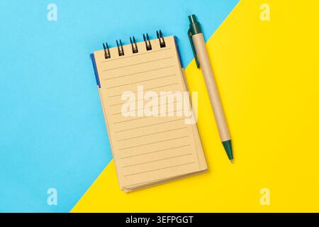 Top view of a spiral notepad with lined pages and an eco-style pen on a blue and yellow background, representing organization, note-taking, planning, and productivity. Stock Photo