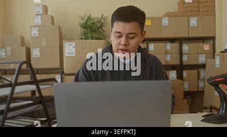 Man typing on laptop in building office surrounded by stacked boxes, shelves and a barcode scanner, wearing a hoodie while checking orders; focused pr Stock Photo