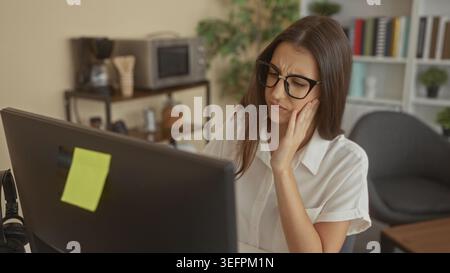 Woman with hand on cheek showing toothache while staring at desktop computer monitor in building; stress. Stock Photo