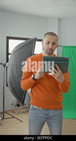 Man holding tablet and gesturing with hand in studio among softbox lights and green screen, wearing orange sweater and jeans; creative concentration. Stock Photo