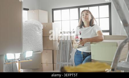 Woman holds packing tape to chin beside stacked cardboard boxes and bubble wrap near ladder in building; quiet contemplation. Stock Photo
