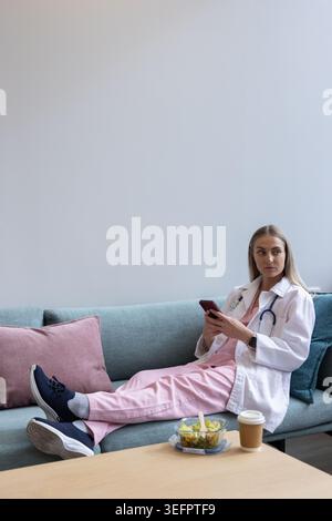 Female clinician reclining on teal sofa in staff room, holding phone, glancing right, copy space Stock Photo