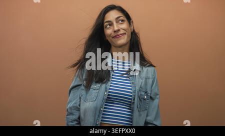 Woman smiling with hair blowing, wearing denim jacket and blue striped crop top in peach studio; contentment. Stock Photo