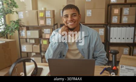 Man with hand on chin at desk with laptop and barcode scanner, surrounded by boxes and shelves in building; small business confidence. Stock Photo