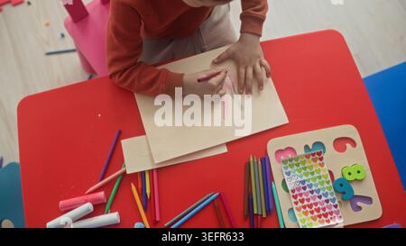 Boy tracing a hand and holding a red crayon while coloring on paper at a red kids table in a studio with stickers and colored pencils; concentration. Stock Photo