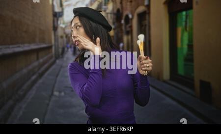 Woman in purple sweater and beret holding paintbrushes, touching cheek with hand in a narrow street; playful curiosity. Stock Photo