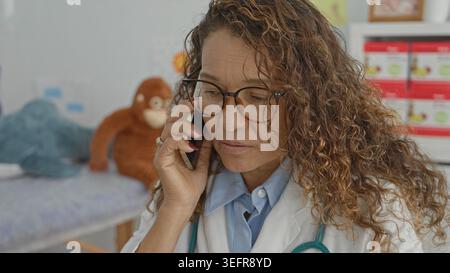 Middle aged woman doctor in white coat holding smartphone to ear, stethoscope visible in clinic exam room with exam bed, plush toy and medical boxes; Stock Photo