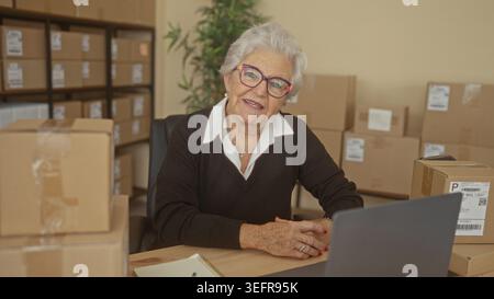 Elderly woman smiling with hands clasped by laptop and packing box in a building small business shipping station, surrounded by stacked parcel boxes; Stock Photo