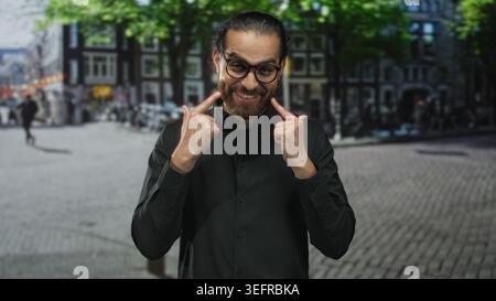 Man wearing glasses and black shirt points index fingers to cheeks and smiles showing teeth on a cobbled city street; joy easy charm. Stock Photo