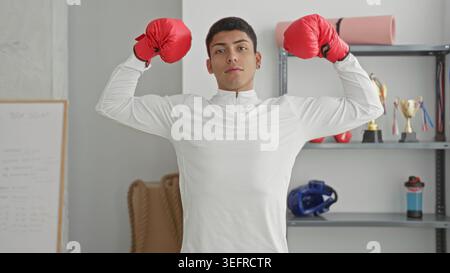 Man young hispanic in white top wearing red boxing gloves flexes biceps and poses in studio among trophies and training gear; confidence discipline. Stock Photo