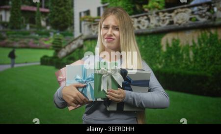 Woman holding wrapped gifts with hands, grimacing while balancing presents in front of a building; frustration burden. Stock Photo