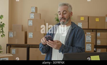 Man holding smartphone and gesturing with hand amid stacked packages and shipping labels in a building; small business joy. Stock Photo
