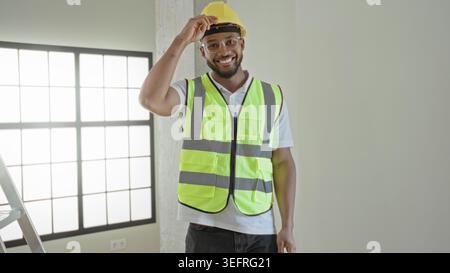 Man tipping hardhat with hand and smiling while wearing reflective vest in a building; confidence professionalism. Stock Photo