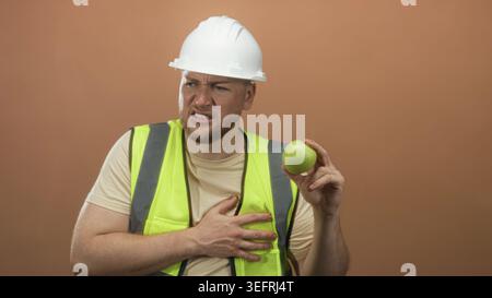 Man engineer in hardhat and reflective vest clutching chest showing heartattack while holding a green apple in a brown studio; concern health safety. Stock Photo