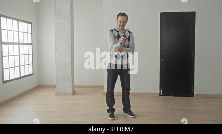 Man holding hammer and inspecting it near window and concrete column in empty house with wooden floor and black door; quiet contemplation planning. Stock Photo