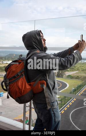 A modern male traveler wearing a hoodie and carrying an orange backpack takes a smartphone selfie from a scenic balcony overlooking a winding road Stock Photo